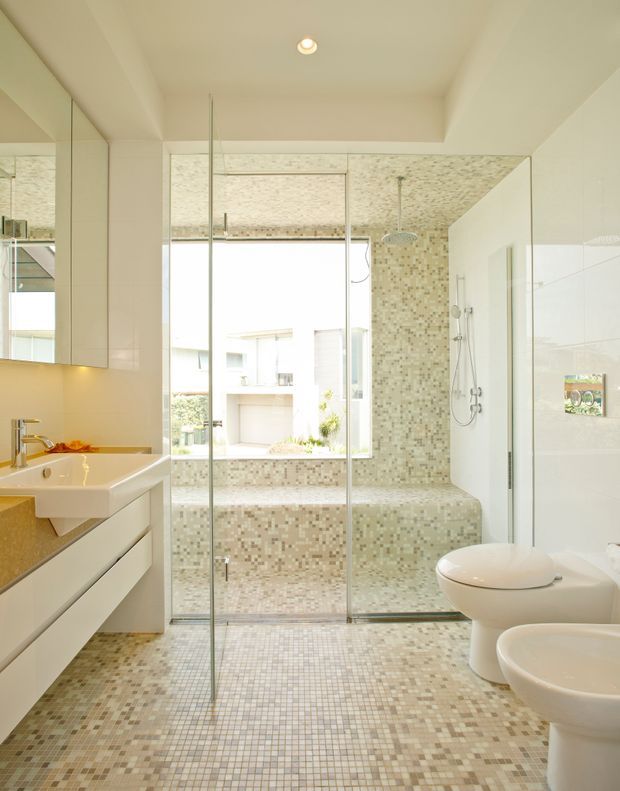 Bright contemporary bathroom with a large frameless glass shower enclosure, built-in tiled bathtub, wall-mounted shower fittings, floating white vanity, vessel sink, toilet and bidet. The room is finished throughout in small beige, cream and grey mosaic tiles, with a large window bringing in abundant daylight and mirrored cabinetry on the left.
