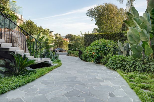 A landscaped exterior walkway paved with irregular grey natural stone, leading through dense tropical and subtropical planting. A concrete staircase with black metal balustrade rises along the left side toward a partially visible building, while hedges and mature garden foliage line the path. Residential buildings and a street are visible in the distance under a bright sky.