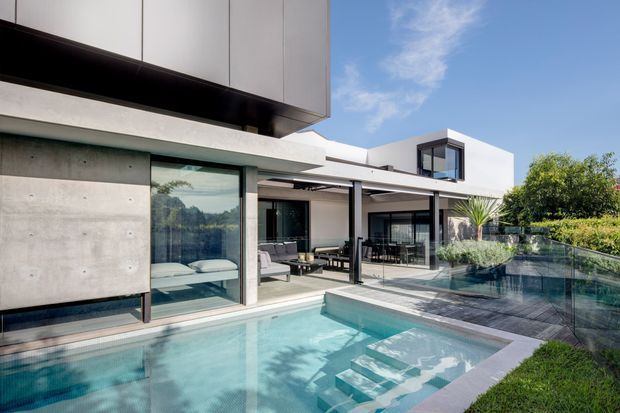 Contemporary multi-level residence with crisp white rendered walls, exposed concrete surfaces, dark metal framing and extensive floor-to-ceiling glazing. The image focuses on a swimming pool and outdoor entertaining terrace adjoining open-plan living spaces, with a sculptural stair descending into the pool and landscaped screening around the property.