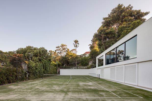 A landscaped residential outdoor tennis court enclosed by dense greenery and rendered boundary walls, beside a modern white, flat-roofed building with extensive dark-framed glazing and louvred panels. The scene has clean geometric lines and soft late-day natural light.