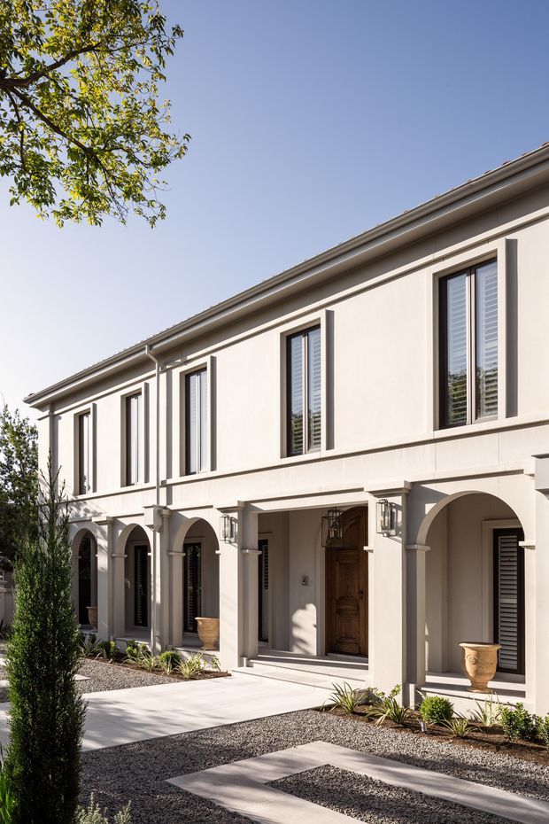 A bright two-storey residential facade with a restrained classical composition. The cream rendered exterior features evenly spaced tall windows with dark frames and shutters, a projecting cornice, and a ground-level colonnade of rounded arches. A timber entry door, traditional wall lanterns, gravel landscaping, clipped planting, and large stone or concrete stepping slabs complete the formal courtyard approach.