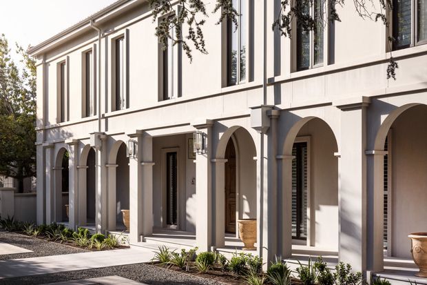 A refined multi-storey residential facade with a pale rendered exterior, repeated arched colonnade, classical pilasters and trim, tall narrow windows, and recessed entry doors. A landscaped gravel forecourt with low planting and decorative urns lines the building.