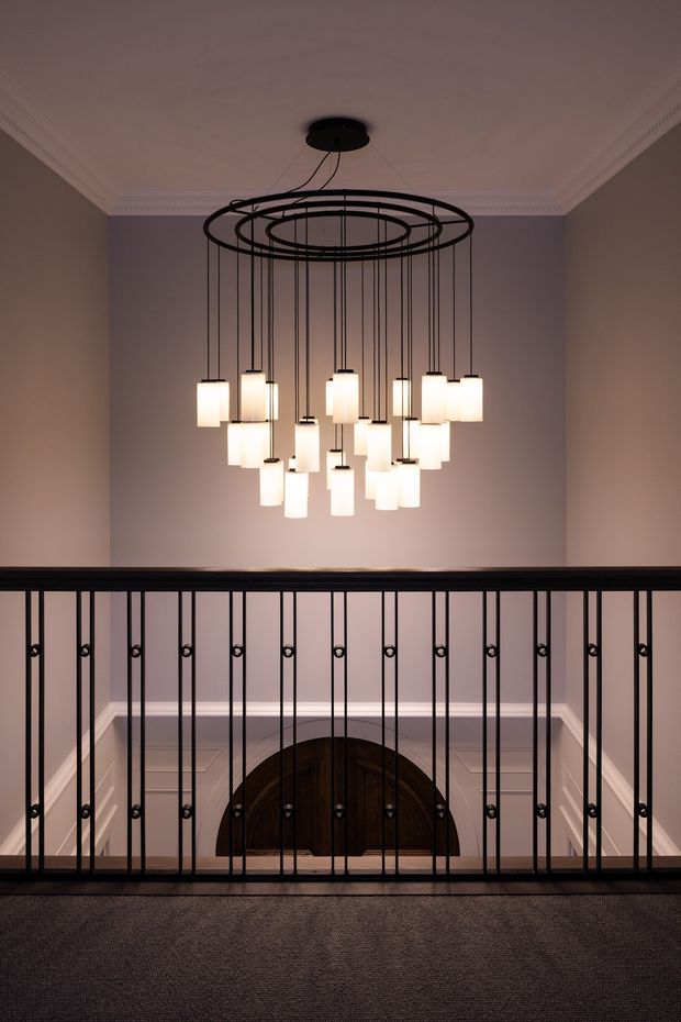 Interior view of an elegant upper-level landing overlooking a staircase, with a large circular black multi-pendant chandelier suspended from a decorative coffered ceiling. The space has muted taupe-grey walls, white ornate cornices and dado-style panelling, dark timber stair elements, black metal balustrading, and carpeted flooring. A rounded dark timber doorway or arched opening is visible below.