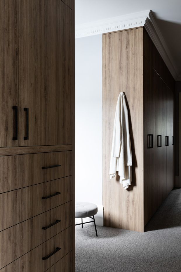 A refined walk-in wardrobe or dressing area with extensive floor-to-ceiling timber-look cabinetry, tall doors, and multiple drawers fitted with slim black handles. A central timber panel holds hanging white garments, while a small upholstered stool sits near a bright window or opening. The space has soft grey carpet, white walls, ornate ceiling cornice detailing, and strong natural light creating a calm, tailored atmosphere.