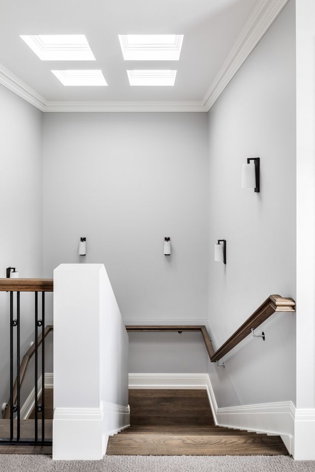 Bright, refined residential stairway viewed from an upper landing, with dark timber stair treads and handrail, white painted balustrade and trim, pale grey walls, black metal balusters, and multiple illuminated rectangular ceiling panels or skylight-like fixtures. The stairwell has traditional moulded cornices and baseboards alongside contemporary minimalist wall sconces.