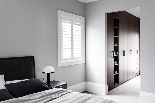 A contemporary bedroom with grey walls, white plantation shutters, a dark upholstered bed, bedside table and lamp, and built-in dark timber wardrobes with open shelving. Soft daylight and neutral textiles create a calm, refined atmosphere.