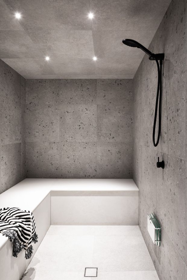 Minimalist walk-in shower with large-format grey stone-look wall tiles, a white tiled floor and built-in white bench. Matte black shower hardware and recessed ceiling spotlights create a clean, monochromatic contemporary bathroom interior. A black-and-white patterned towel adds a small decorative accent.