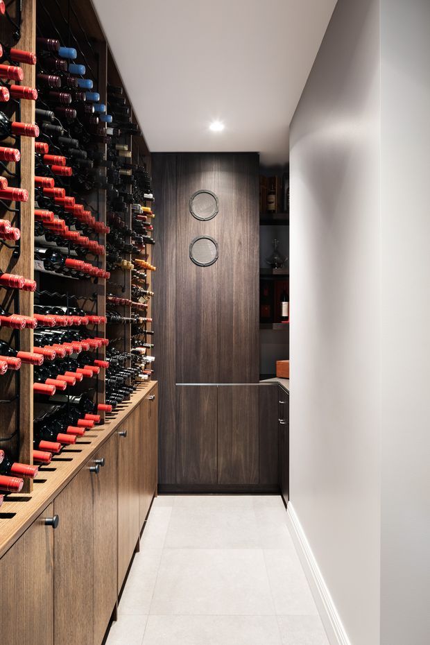 A narrow, contemporary wine storage room with extensive wall-mounted bottle racks on the left, dark timber cabinetry and a tall central cabinet with two circular inset details. Pale large-format floor tiles and white walls create contrast with the dark wood joinery, while recessed ceiling lighting provides focused illumination. Additional bottle storage and shelving are visible at the rear.