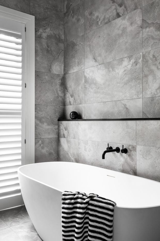 A refined monochromatic bathroom featuring a freestanding white bathtub against large-format grey stone-look wall tiles. A matte black wall-mounted bath tap and slim black shelf provide contrast, while white plantation shutters bring in filtered light. A striped black-and-white towel hangs over the tub.