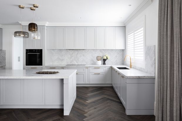 Bright, refined white kitchen with a large central island, U-shaped perimeter cabinetry, marble-look backsplash and benchtops, integrated black oven, brass tapware, pendant lights, plantation shutters, and dark herringbone timber flooring. The space has a polished modern-classic appearance with strong symmetry and abundant natural light.