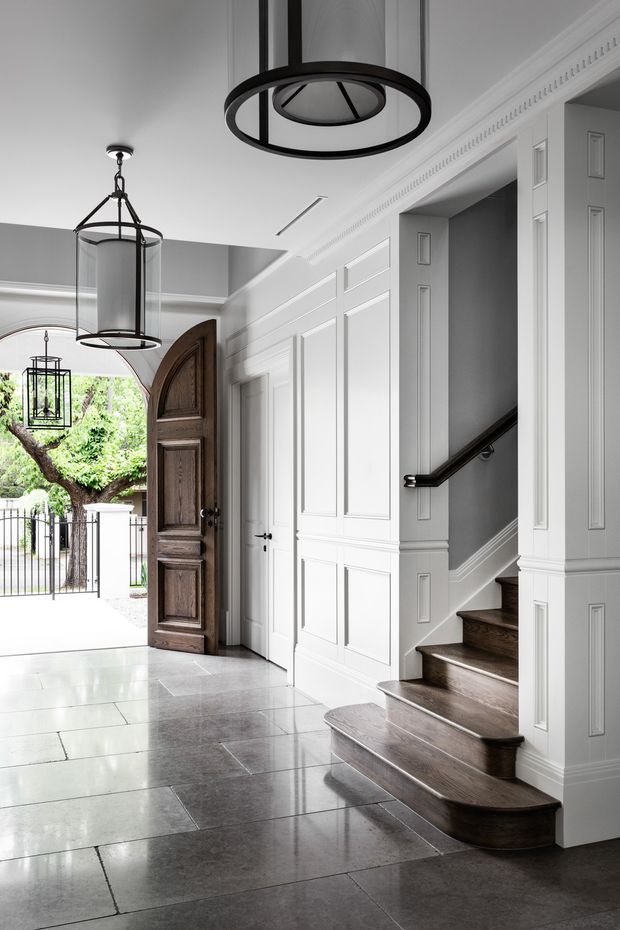 A refined residential entry foyer with a highly detailed white paneled interior, ornate crown moulding, dark timber front door, and a staircase with curved dark wood treads and black handrail. Large grey stone floor tiles and black-framed pendant lights create a monochrome, elegant composition. The open doorway leads outside to a garden, but the foyer and staircase dominate the image.