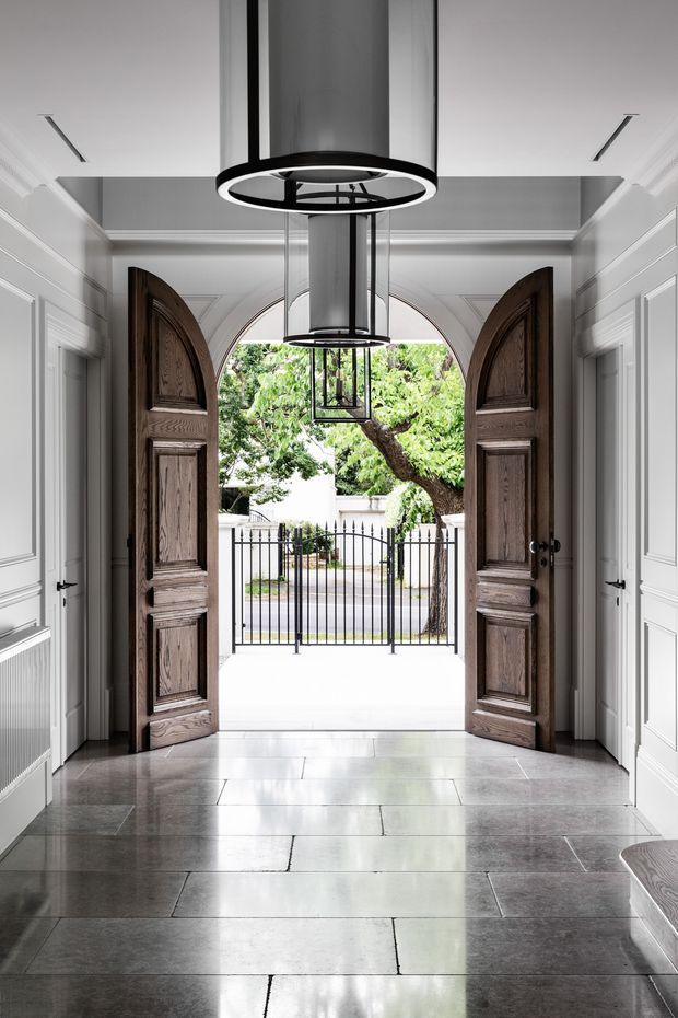 Formal residential entry hall viewed from inside toward tall, open, dark timber double doors. The space features grey stone slab flooring, white painted walls with traditional raised panel moulding, arched architectural detailing around the doorway, and large black cylindrical pendant lights. A gated exterior forecourt and leafy tree are visible beyond the entrance.