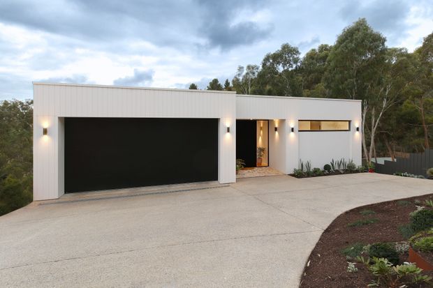 A minimalist modern single-storey home exterior with a white vertical-panel facade, broad flat roofline, large black garage door, recessed illuminated entry, and narrow horizontal window. A broad concrete driveway and landscaped garden bed lead up to the house, with eucalyptus trees in the background under an overcast sky.