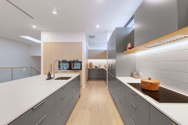 A bright contemporary kitchen with a long central island, extensive grey built-in cabinetry, white benchtops, integrated appliances, and a tiled cooking backsplash. Pale timber flooring and timber accents add warmth, while skylights and recessed lighting create an airy feel. A stair balustrade is visible at the left.