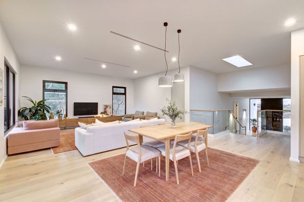 Bright open-plan living and dining area with pale timber flooring, a large light timber dining table and chairs, sofas, a television, pendant lights, skylight, and expansive windows. The space opens toward a stair and lower-level area, with a warm neutral palette and contemporary furnishings.