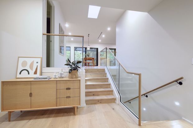 Bright contemporary stairway and landing with pale timber flooring and matching timber stair treads, a glass balustrade with slim timber handrails, white walls, recessed lighting and a skylight. A mid-century-inspired timber sideboard and artwork occupy the landing, with a dining area visible beyond.