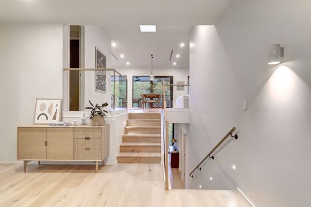 Bright contemporary stair landing and hallway with pale timber flooring, white walls, recessed ceiling lights, and a warm timber staircase descending to a lower level. A low timber sideboard, framed artwork, handrail, and glimpses of a dining area and outdoor greenery are visible.
