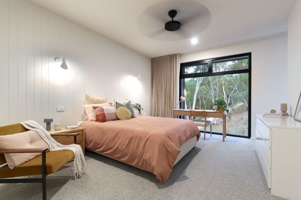 A warm, contemporary bedroom with a soft peach-pink upholstered bed, layered cushions and throws, pale vertical wall panelling, grey carpet, and a mustard lounge chair. Black-framed floor-to-ceiling glazing opens to a leafy outlook, while a timber desk and white dresser provide practical storage and workspace. Soft pendant and wall lighting, along with neutral curtains, create a calm and inviting atmosphere.