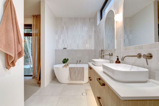 Bright contemporary bathroom with a freestanding white bathtub, double vessel basins on a timber vanity, brushed metal wall-mounted tapware, large arched mirrors, and pale textured wall tiles. A doorway reveals an adjacent carpeted room and hanging towels.