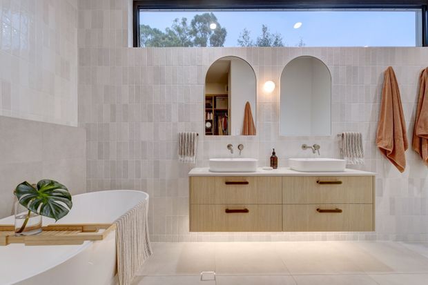 Bright contemporary bathroom with a floating light timber double vanity, two vessel basins, arched recessed niches and mirrors, pale vertically patterned wall tiles, and a freestanding bathtub partially visible at left. A high horizontal window provides natural light, while warm towels and subtle styling add softness.