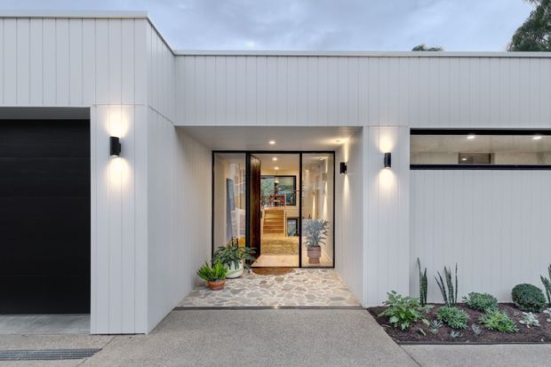 Street-facing view of a contemporary single-storey residence with a white vertically panelled facade, flat rooflines, black garage door, recessed glazed entry, exterior wall lights, and a paved front path bordered by low planting. Warm interior timber finishes are visible through the entry glazing.