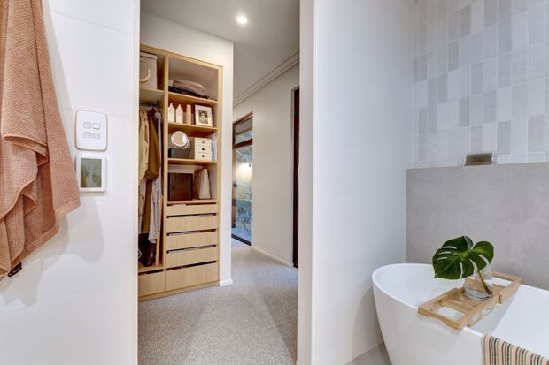 A bright, refined bathroom viewed from the entry, with a freestanding white bathtub in the foreground, a pale tiled feature wall, and a timber bath tray holding a plant and accessories. Beyond the bathroom is a carpeted dressing area with open timber wardrobe cabinetry, drawers, hanging clothes, and shelving. White walls, recessed lighting, and a neutral palette create a clean, calm atmosphere.
