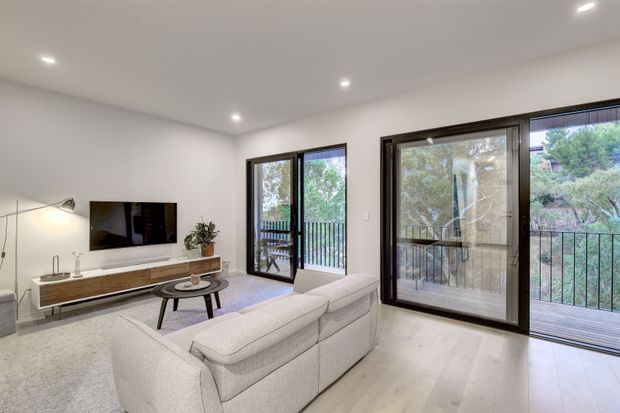 Bright contemporary living room with a neutral palette, pale timber flooring, light upholstered seating, a low timber media console, recessed ceiling lights, and expansive dark-framed glazed doors opening to a balcony or terrace overlooking trees.
