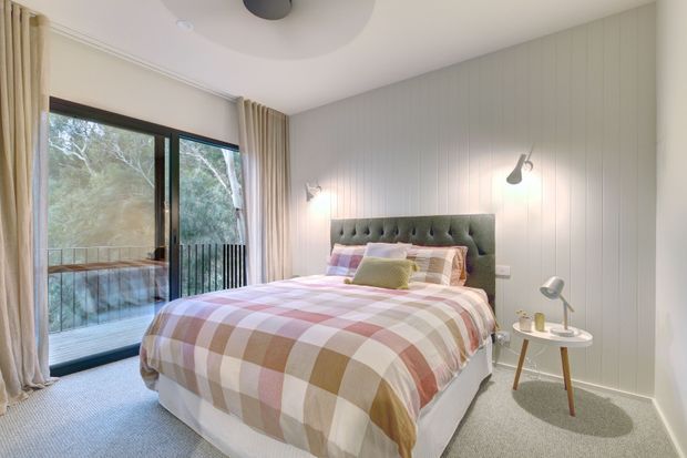 A bright, cosy bedroom with a tufted upholstered bedhead, layered checked and pastel bedding, pale vertical wall panelling, soft carpet, bedside tables, wall-mounted reading lights, and full-height glazed doors opening to a balcony or exterior view. Neutral curtains and abundant natural light create a relaxed contemporary retreat.