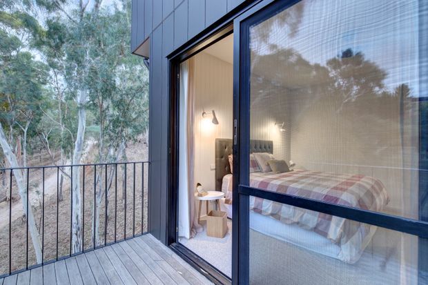Exterior view of a contemporary bedroom opening onto a timber-floored balcony or terrace, surrounded by trees. The room is visible through large dark-framed glazed doors and includes a bed, upholstered headboard, bedside table, and wall-mounted reading light. A screened sliding door is partly closed in the foreground.