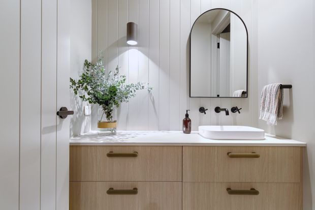 A bright, refined bathroom featuring a floating light-wood vanity with four drawers, a white vessel basin, wall-mounted matte-black tapware, a rounded-arch mirror, white vertical wall panelling, and a minimal wall sconce. A towel rail and small decorative accessories complete the clean, warm scheme.