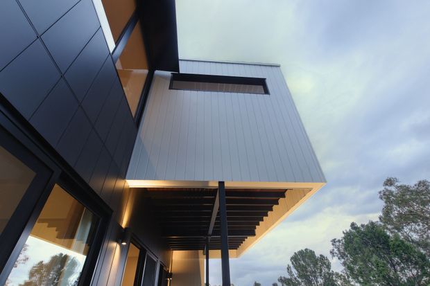 Upward-looking exterior architectural photograph of a contemporary building, showing a projecting upper-level volume clad in light vertical panels, dark metal wall panels and window frames, a deep cantilevered eave with exposed dark rafters, and warm recessed lighting along the covered outdoor walkway. Trees and overcast sky are visible beyond the structure.