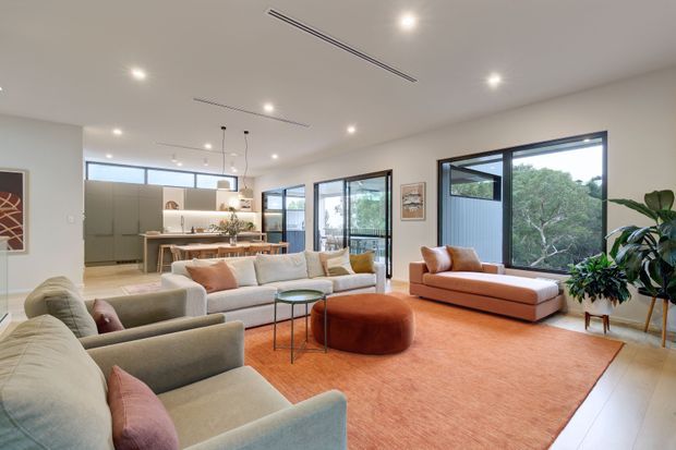 Bright, spacious contemporary open-plan living room with large neutral sofas, a rust-orange rug and ottoman, and a matching low-profile lounge. Floor-to-ceiling glazing opens views to greenery, while a modern kitchen and dining area are visible in the background. Recessed ceiling lights and linear air vents contribute to the clean architectural finish.