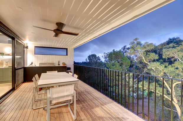 Covered outdoor entertaining area with a timber deck, dining table and chairs, built-in bar cabinetry, ceiling fan, and a black metal balustrade overlooking leafy treetops. The space connects to the interior through large glazed sliding doors and features a white-lined ceiling with integrated lighting.
