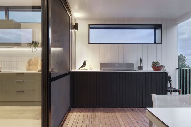 Contemporary open-plan interior showing a dark vertically slatted kitchen or bar island, pale timber wall panelling, a horizontal clerestory-style window, and warm timber flooring. A light-toned kitchen is visible through a glazed or open partition on the left, while a dining table occupies the foreground. The space has clean lines, restrained decor, and views toward greenery outside.