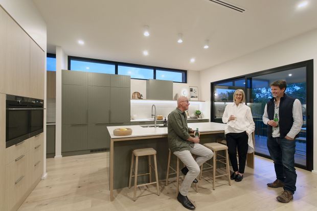 Bright contemporary open-plan kitchen with a large central island, integrated light-wood cabinetry, muted green-grey pantry cabinetry, white benchtops and a wall of dark-framed glazing opening to the outdoors. The island has timber stools and casual seating, while several people stand and sit in the space. Recessed ceiling lights and pale timber flooring create an airy, welcoming interior.