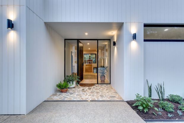 Contemporary residential entrance viewed from outside, featuring a recessed porch clad in vertical white panels, black-framed glazed entry doors, a pebble-patterned stone floor, integrated wall lighting, and landscaped planting beds. The interior hallway and timber joinery are visible through the glazing.