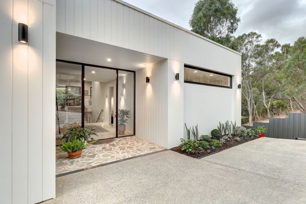 Modern single-storey residential facade with white vertical cladding, a flat roofline, dark-framed glazed entry doors, and a narrow horizontal window. A patterned stone entry path, exposed aggregate driveway, wall lighting, and minimal planted beds complete the clean contemporary exterior.