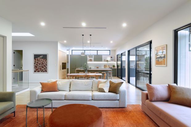 Bright open-plan contemporary living room with a large light upholstered sectional sofa, warm rust-orange cushions and rug, rounded coffee table, and extensive black-framed glazing. A dining area and modern kitchen are visible beyond, with pale walls, timber cabinetry, and linear pendant lighting.