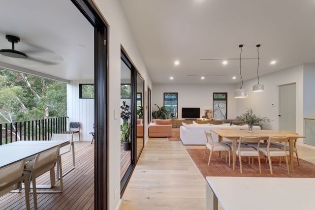 Bright open-plan contemporary living, dining and kitchen area with pale timber flooring, white walls and ceiling, large black-framed glazed doors, and a covered timber deck opening to leafy surroundings. The living zone includes a pale sofa and television, while the dining area has a timber table and chairs beneath simple pendant lights. A ceiling fan and outdoor dining furniture are visible on the deck.