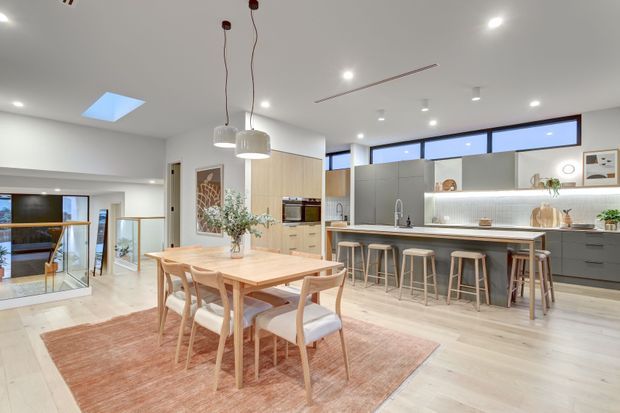 Bright open-plan kitchen and dining area with a light timber dining table and chairs on a muted terracotta rug, facing a contemporary grey kitchen with a long island, timber benchtop, integrated appliances, pendant lights, and multiple stools. A glazed stair landing and living area are visible beyond, with skylights and clerestory-style windows adding natural light.