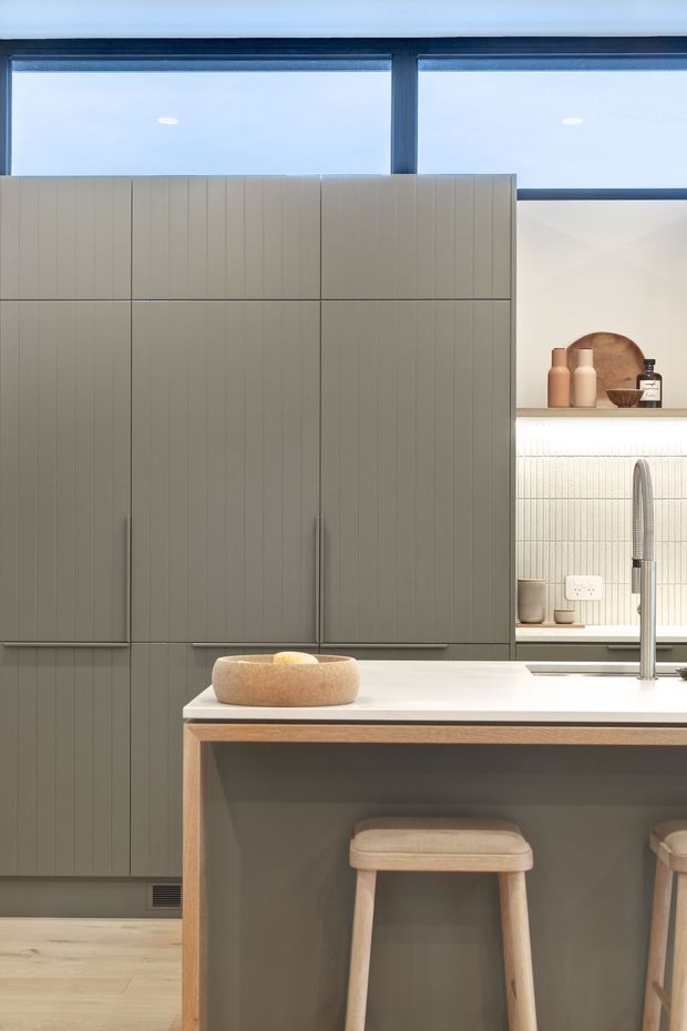 A refined contemporary kitchen featuring a large central island with a white benchtop, integrated seating, and a tall brushed-metal tap. Full-height muted grey-green cabinetry has subtle vertical fluting and concealed handles. Behind the island is a softly illuminated ribbed tile splashback with open display shelving, neutral ceramics, and warm-toned vessels. Large upper windows bring in natural light, while pale timber flooring and warm timber stool details soften the restrained palette.