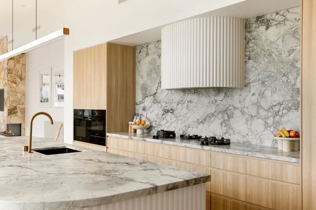 A bright, refined contemporary kitchen featuring a large marble island with an integrated dark sink and a brass gooseneck tap. The rear wall combines full-height veined marble splashback with light timber cabinetry, black integrated appliances, a long timber drawer bank, and a sculptural white fluted rangehood. A linear pendant light and subtle display shelving complete the clean, warm-toned space.