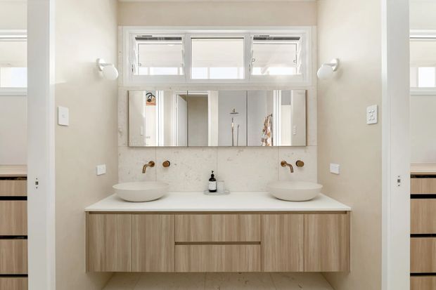 Bright, symmetrical contemporary bathroom vanity with two round countertop basins, wall-mounted warm metallic taps, pale stone or porcelain benchtop, and a floating timber-look double vanity. A wide mirrored cabinet sits beneath three high horizontal windows, with wall sconces on either side. Partial adjoining cabinetry is visible through the side openings.