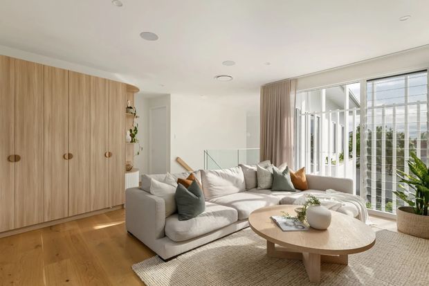 Bright, contemporary living room with a large neutral sectional sofa, round light-wood coffee table, textured area rug, built-in timber cabinetry, indoor plants, and full-height glazing with vertical privacy screens. A stair landing is visible beyond the seating area. The space has a warm, airy, refined atmosphere with abundant natural light.