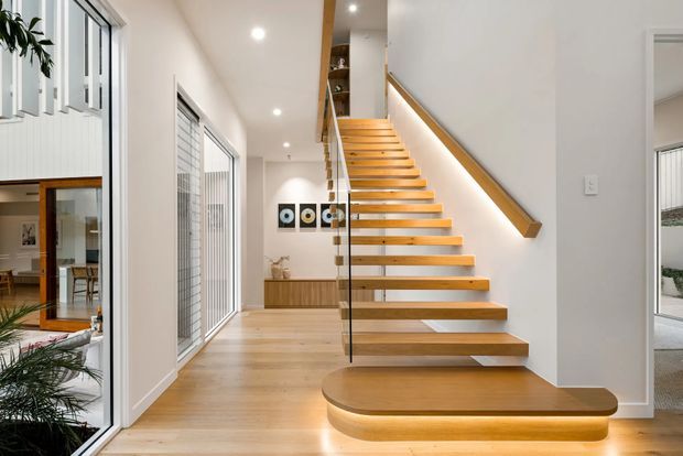 Bright contemporary interior hallway centred on a sculptural open-riser timber staircase. The stairs feature thick light-to-mid timber treads, a rounded platform step, frameless glass balustrade, and a timber handrail with concealed under-step lighting. White walls, recessed ceiling lights, pale timber flooring, and large glazed openings create an airy, refined entry circulation space. A small glimpse of a kitchen or living area is visible beyond the stair.
