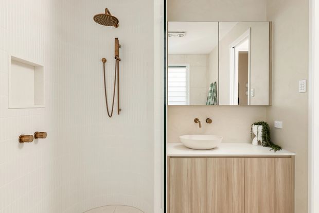 Contemporary bathroom featuring a walk-in shower with white vertically textured wall tiles, brushed brass shower fittings and recessed niche, alongside a floating timber-look vanity with a white vessel basin, mirrored cabinet and brass wall-mounted tapware. Neutral beige and white palette with subtle greenery.
