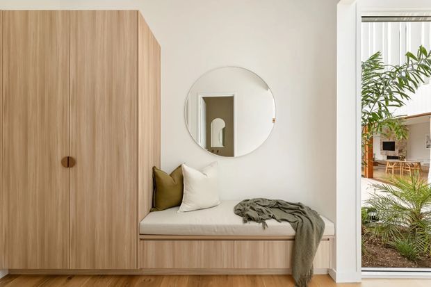 A bright, softly styled entryway or compact living nook featuring a full-height light timber built-in cabinet, an integrated upholstered bench seat with cushions and throw, a round wall mirror, and a large glazed opening overlooking planted greenery and an adjacent interior. The space has a clean, contemporary minimalist aesthetic with warm natural materials.