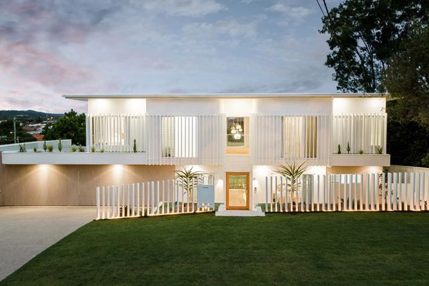 Twilight exterior view of a contemporary two-storey residence with a long, low flat roof and strong vertical white screening across the facade. Warm interior and facade lighting highlights recessed balconies, glazing, entry doors, a covered carport or driveway area, landscaped planters, and a broad lawn. The design has a crisp geometric composition with white rendered surfaces and vertical slatted elements.