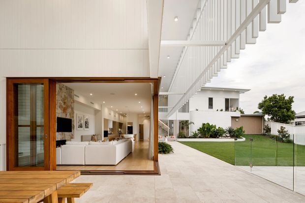 Contemporary residential courtyard view showing a bright open-plan living area opening directly onto a spacious pale-paved terrace and manicured lawn. The interior features a white sectional sofa, timber flooring and framing, a textured stone feature wall, recessed lighting and a kitchen beyond. A large white cantilevered or projecting upper structure with vertical screening elements extends over the terrace, while frameless glass balustrading encloses the outdoor edge.