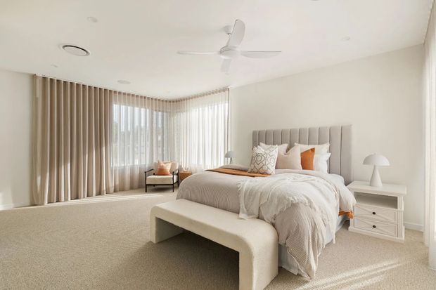 Bright, spacious contemporary bedroom with a large upholstered bed, soft neutral bedding, a curved upholstered bench, white bedside tables, and layered floor-to-ceiling curtains around corner windows. A white ceiling fan, recessed lighting, and a small reading chair complete the calm, refined interior.